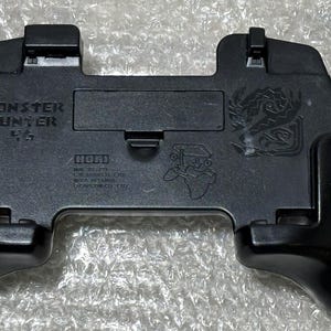 Może przedstawiać: Czarny kontroler do gier z napisem "MONSTER HUNTER" i "4G". Kontroler ma teksturowaną powierzchnię i ergonomiczne uchwyty. Stylizowana grafika smoka jest widoczna po prawej stronie u góry. Marka "HORI" jest również wydrukowana.