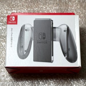 Może przedstawiać: Uchwyt ładujący Joy-Con do Nintendo Switch w oryginalnym opakowaniu. Pudełko jest czerwono-białe, z widocznym logo Nintendo Switch. Uchwyt jest szary i przezroczysty, przeznaczony do kontrolerów Joy-Con.
