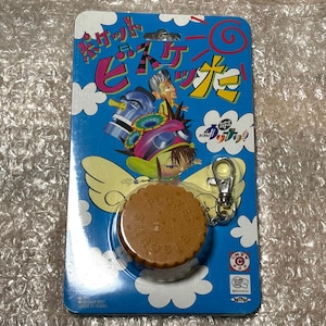 Puede incluir: Un llavero con un dije en forma de galleta marrón con el texto "ROCKET" y "SMILE" en él. El llavero está unido a una cadena de metal plateado con un anillo de metal plateado. El empaque presenta una ilustración de dibujos animados de un personaje con una galleta en la cabeza y el texto "ROCKET SMILE".