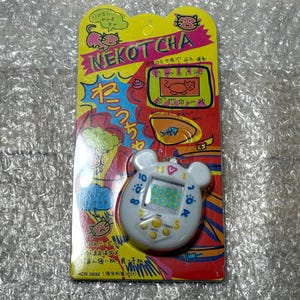 May include: A white plastic cat-shaped electronic pet toy with a screen that displays a cat face and the text "NEKOT CHA" on the packaging. The packaging is pink, yellow, and blue with Japanese text.