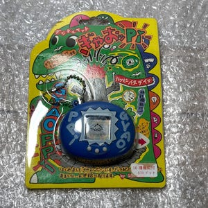 K&ouml;nnte beinhalten: Ein blauer und wei&szlig;er digitaler Haustier-Schl&uuml;sselanh&auml;nger mit einem gr&uuml;nen Dinosaurier und dem Text "Happy Birthday" auf der Verpackung. Der Schl&uuml;sselanh&auml;nger ist mit "570 Punkten" gekennzeichnet.