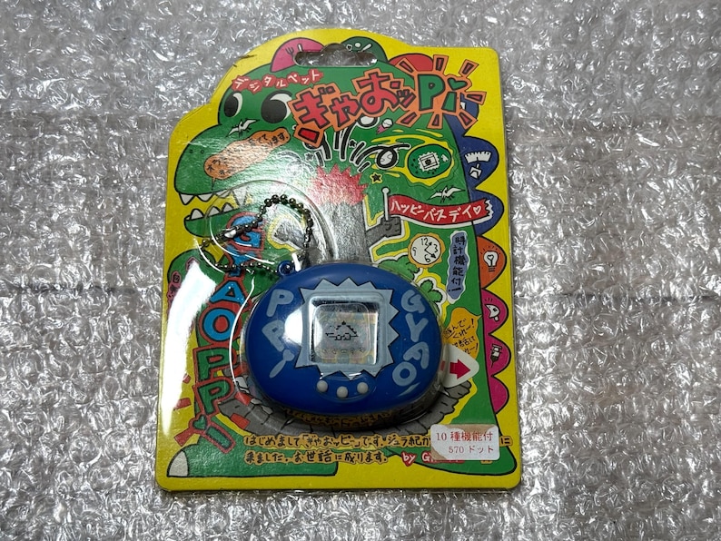 K&ouml;nnte beinhalten: Ein blauer und wei&szlig;er digitaler Haustier-Schl&uuml;sselanh&auml;nger mit einem gr&uuml;nen Dinosaurier und dem Text "Happy Birthday" auf der Verpackung. Der Schl&uuml;sselanh&auml;nger ist mit "570 Punkten" gekennzeichnet.