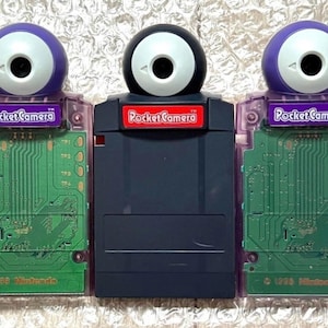 Può includere: Tre dispositivi Pocket Camera vintage. Due hanno teste di fotocamera viola e uno ha una testa di fotocamera nera. Le cartucce sono viola scuro e grigio scuro, con circuiti stampati verdi visibili. Il testo "Pocket Camera" è visibile.