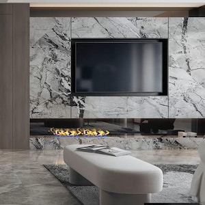 Puede incluir: Un banco blanco moderno con una parte superior curva y dos patas cilíndricas se encuentra frente a una chimenea con un televisor negro montado encima. La chimenea está rodeada por una pared de mármol gris con vetas blancas.