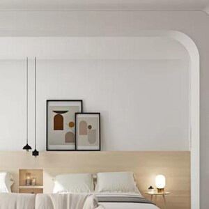 Puede incluir: Un dormitorio moderno con una cabecera blanca y una puerta arqueada blanca. El dormitorio tiene dos mesitas de noche con lámparas y una cama con ropa de cama blanca. Hay dos impresiones abstractas enmarcadas en la pared sobre la cabecera.