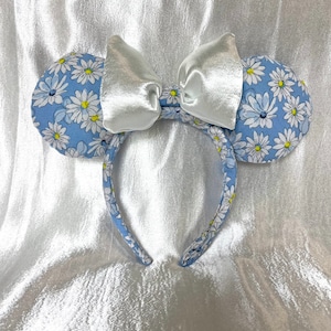 May include: A pair of blue and white floral Minnie Mouse ears with a white satin bow. The ears are made of fabric and have a headband.