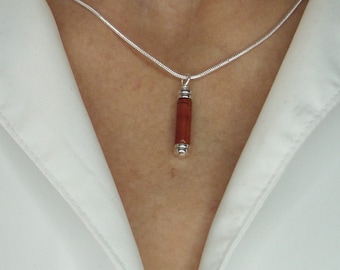 Può includere: Una collana d'argento con un pendente cilindrico, rosso-arancio. Il pendente è rifinito con cappucci argentati su ogni estremità. La collana è indossata sopra una camicia bianca.