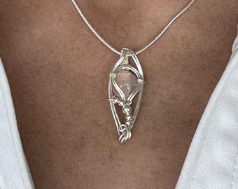 May include: A silver pendant necklace featuring a light pink gemstone encased in a wire-wrapped design. The pendant hangs from a delicate silver chain. The necklace is displayed against a white shirt.
