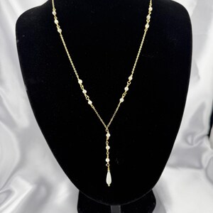 Pu&ograve; includere: Una collana a catena color oro con piccole perle bianche distanziate uniformemente lungo la catena. La collana presenta un design a Y che termina con un singolo ciondolo a forma di perla bianca allungata a goccia al centro.