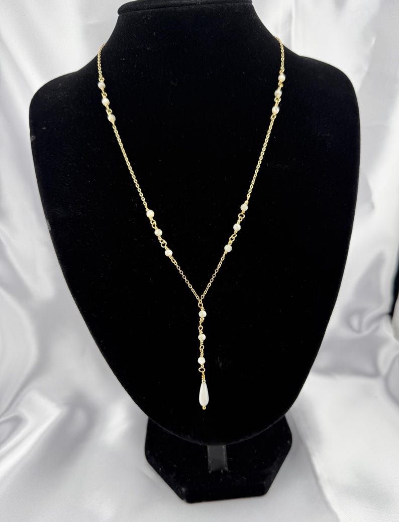 Pu&ograve; includere: Una collana a catena color oro con piccole perle bianche distanziate uniformemente lungo la catena. La collana presenta un design a Y che termina con un singolo ciondolo a forma di perla bianca allungata a goccia al centro.