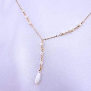 Pu&ograve; includere: Una collana dorata a forma di Y caratterizzata da una catenina sottile decorata con piccole perline rotonde color bianco sporco distanziate lungo la catena, che termina con una singola perla bianca allungata a goccia al centro.