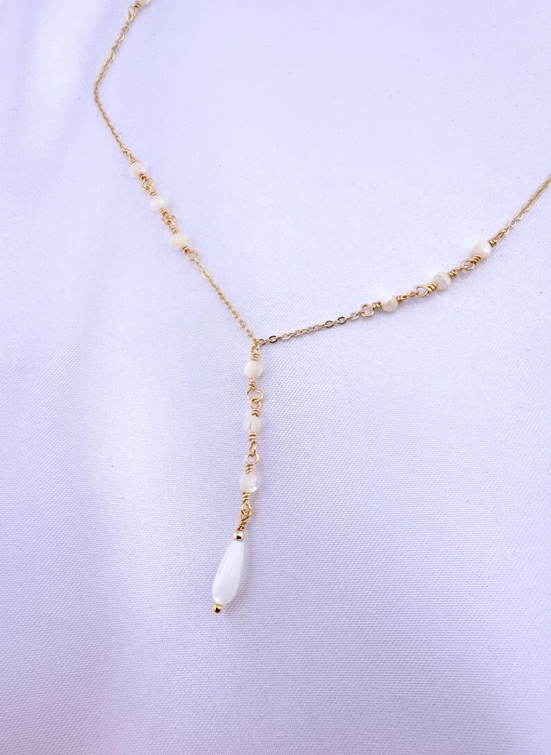 Pu&ograve; includere: Una collana dorata a forma di Y caratterizzata da una catenina sottile decorata con piccole perline rotonde color bianco sporco distanziate lungo la catena, che termina con una singola perla bianca allungata a goccia al centro.
