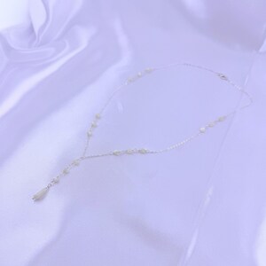 Pu&ograve; includere: Una delicata collana a catena color argento con piccole perline rotonde color bianco sporco distanziate lungo la catena. La collana ha un design a Y, che termina con una singola perla allungata a forma di goccia al centro.