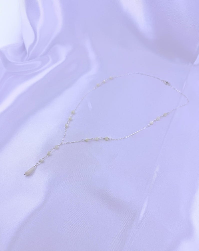 Pu&ograve; includere: Una delicata collana a catena color argento con piccole perline rotonde color bianco sporco distanziate lungo la catena. La collana ha un design a Y, che termina con una singola perla allungata a forma di goccia al centro.