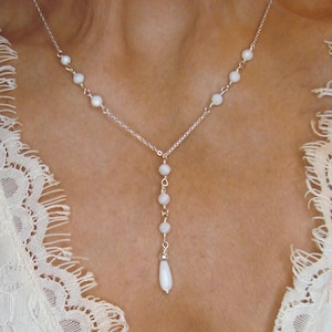 Pu&ograve; includere: Una collana a catena color argento con piccole perline bianche rotonde distanziate lungo la catena, con una sezione centrale verticale che termina con un ciondolo a forma di goccia bianca allungata.