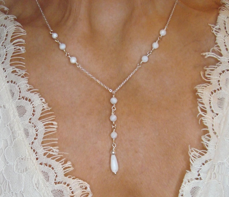 Pu&ograve; includere: Una collana a catena color argento con piccole perline bianche rotonde distanziate lungo la catena, con una sezione centrale verticale che termina con un ciondolo a forma di goccia bianca allungata.