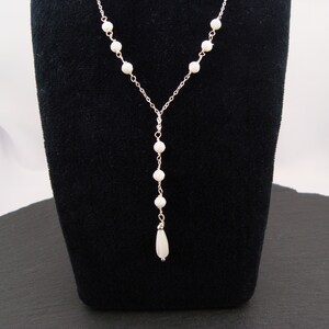 Pu&ograve; includere: Una collana a forma di Y color argento con una catenina sottile decorata con piccole perline bianche rotonde. La collana termina con un unico ciondolo a forma di goccia bianca allungata, sospeso al centro della struttura della catena.