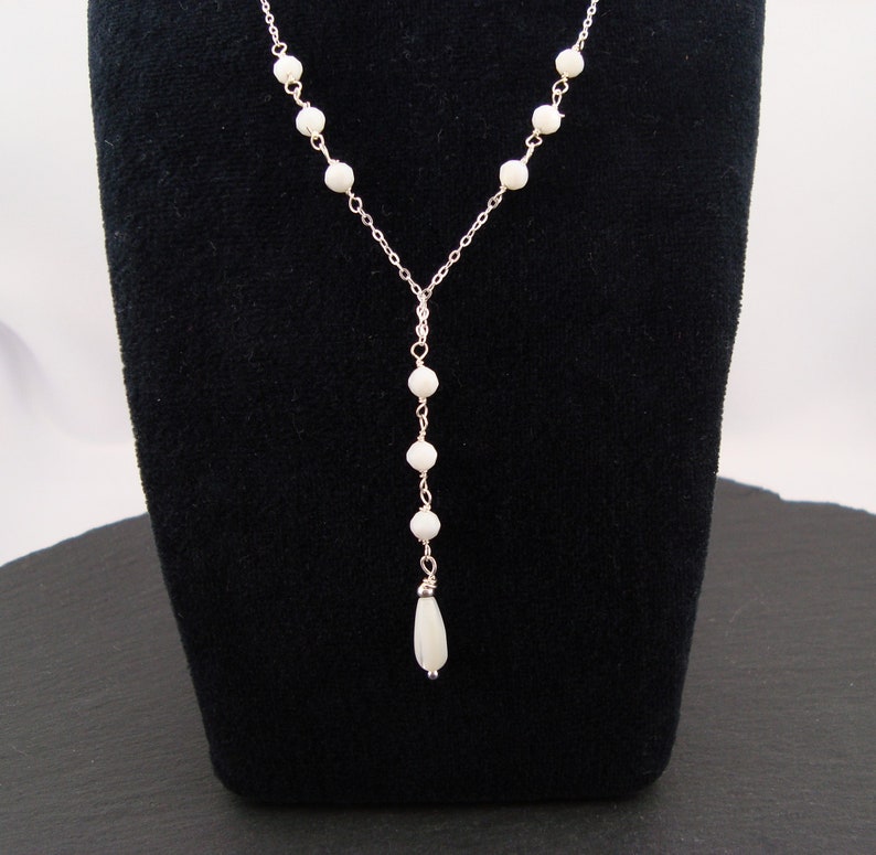 Pu&ograve; includere: Una collana a forma di Y color argento con una catenina sottile decorata con piccole perline bianche rotonde. La collana termina con un unico ciondolo a forma di goccia bianca allungata, sospeso al centro della struttura della catena.