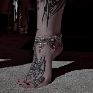 Op de afbeelding: Een zilveren maliënkolder enkelband met een decoratieve franje hangt aan een enkel. De voet heeft een zwart-witte kasteeltattoo. De teennagels zijn zwart gelakt. De foto is binnenshuis genomen.