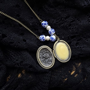 Könnte beinhalten: Eine Halskette mit einem Medaillon im Vintage-Stil mit einer Bronzekette und einem Anhänger. Das offene Medaillon zeigt einen gelben Innenraum. Die Kette hat blaue und weiße Blumenperlen. Das Medaillon hat ein Blumendesign auf der Vorderseite.