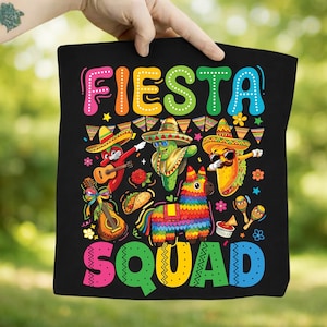 May include: Black tote bag featuring the words "Fiesta Squad" in colorful bubble letters. The design includes cartoon characters of a chili pepper, cactus, and taco wearing sombreros, with a guitar, maracas, and a pinata.