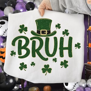 May include: White sweatshirt with a green graphic that says "BRUH" with a green leprechaun hat and shamrocks. The sweatshirt is held up by a person. The background has Halloween decorations.