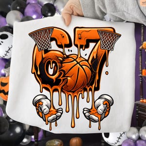 May include: White sweatshirt featuring a basketball-themed graphic. The design includes the number 67, a basketball, and two basketball hoops, all in shades of orange and black. The graphic has a dripping effect, with cartoon hands at the bottom.