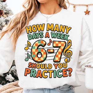 May include: White sweatshirt with a colorful graphic. The graphic reads "HOW MANY DAYS A WEEK 6-7 SHOULD YOU PRACTICE?" with musical notes and a treble clef. The numbers 6 and 7 have a checkered pattern.