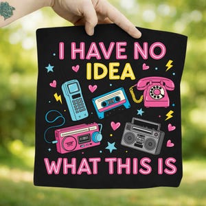 Puede incluir: Bolso tote negro con un gráfico colorido de electrónica retro, incluyendo un teléfono, una cinta de cassette y una radio. El texto dice "I HAVE NO IDEA WHAT THIS IS" en rosa y amarillo.