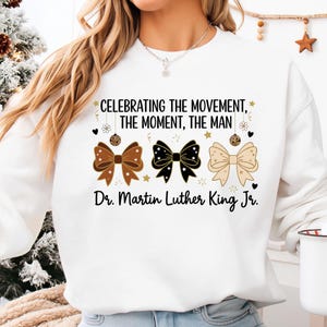 May include: White sweatshirt featuring three decorative bows in brown, black, and beige, with star accents. The text "CELEBRATING THE MOVEMENT, THE MOMENT, THE MAN" and "Dr. Martin Luther King Jr." are printed on the front.