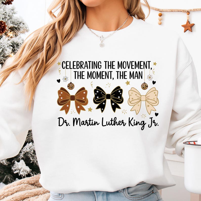 May include: White sweatshirt featuring three decorative bows in brown, black, and beige, with star accents. The text "CELEBRATING THE MOVEMENT, THE MOMENT, THE MAN" and "Dr. Martin Luther King Jr." are printed on the front.