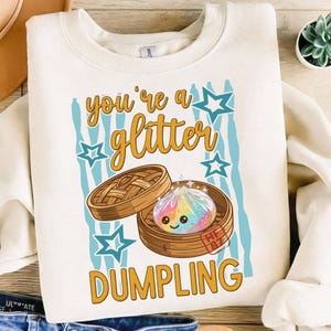 May include: Cream-colored sweatshirt with a graphic featuring a smiling, rainbow-colored dumpling in a bamboo steamer. The text "you're a glitter DUMPLING" is above and below the dumpling, with blue star and stripe accents.