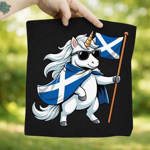 May include: Black tote bag featuring a white unicorn wearing sunglasses and a blue and white Scottish flag cape. The unicorn holds a Scottish flag on a wooden pole. The design is a cartoon illustration.