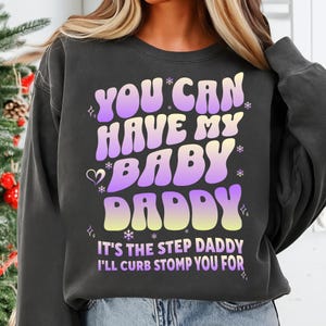 May include: Dark gray sweatshirt with the text "YOU CAN HAVE MY BABY DADDY" in a gradient purple and yellow font. The phrase "IT'S THE STEP DADDY I'LL CURB STOMP YOU FOR" is printed below the main text.