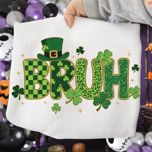 May include: White sweatshirt with the word "BRUH" in green, featuring a checkered pattern, leopard print, and solid green letters. A green leprechaun hat and shamrocks are above and around the word. Gold stars are scattered around the design.