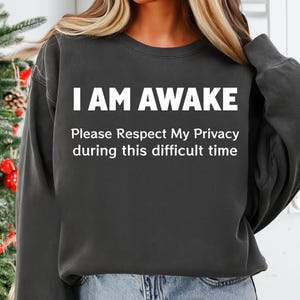 Peut inclure: Sweat-shirt gris foncé avec l'inscription blanche "I AM AWAKE". En dessous, un texte blanc plus petit indique "Please Respect My Privacy during this difficult time."