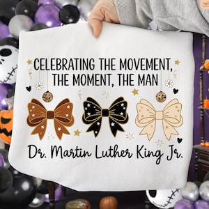 May include: White sweatshirt with the text "CELEBRATING THE MOVEMENT, THE MOMENT, THE MAN" and "Dr. Martin Luther King Jr." in black. The design includes three bow ties in brown, black, and beige, with stars and disco ball accents.