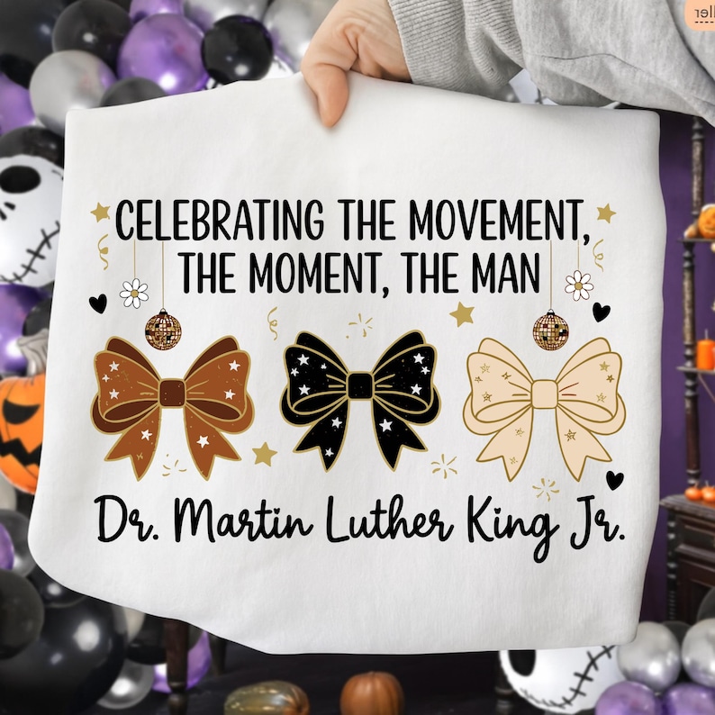 May include: White sweatshirt with the text "CELEBRATING THE MOVEMENT, THE MOMENT, THE MAN" and "Dr. Martin Luther King Jr." in black. The design includes three bow ties in brown, black, and beige, with stars and disco ball accents.