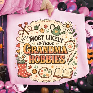 May include: A light pink sweatshirt with the text "MOST LIKELY to Have GRANDMA HOBBIES" in orange and brown. The design includes illustrations of knitting needles, flowers, a watering can, and other craft-related items.