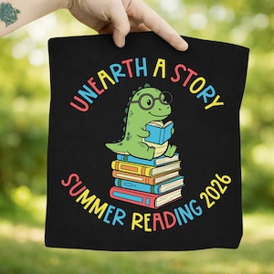 Könnte beinhalten: Schwarze Tragetasche mit einem grünen Dinosaurier mit Brille, der ein Buch liest und auf einem Stapel Bücher sitzt. Der Text "UNEARTH A STORY SUMMER READING 2026" ist in einem farbenfrohen Bogen um den Dinosaurier gedruckt.