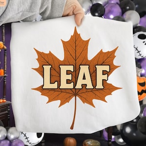 Peut inclure: Taie d'oreiller blanche avec une grande feuille d'érable orange détaillée. Le mot "LEAF" est imprimé dans une police crème audacieuse à l'intérieur de la feuille. La taie d'oreiller est tenue par une personne.