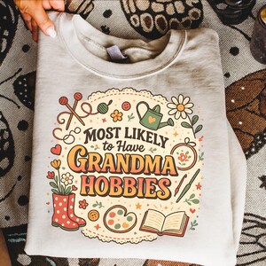 May include: A light gray sweatshirt with the text "MOST LIKELY to Have GRANDMA HOBBIES" in orange and yellow. The design includes illustrations of knitting needles, flowers, a watering can, and other craft-related items.