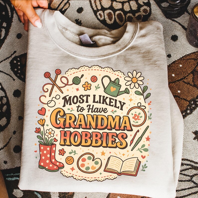 May include: A light gray sweatshirt with the text "MOST LIKELY to Have GRANDMA HOBBIES" in orange and yellow. The design includes illustrations of knitting needles, flowers, a watering can, and other craft-related items.