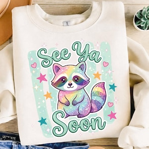Puede incluir: Sudadera crema con un gráfico de mapache colorido. El mapache tiene pelaje arcoíris y está rodeado de estrellas y corazones. El texto "See Ya Soon" está arriba y abajo del mapache.