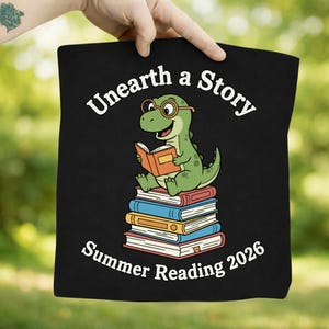 Può includere: Borsa tote nera con un dinosauro verde con gli occhiali che legge un libro, seduto su una pila di libri. Il testo "Unearth a Story" e "Summer Reading 2026" è stampato in bianco.