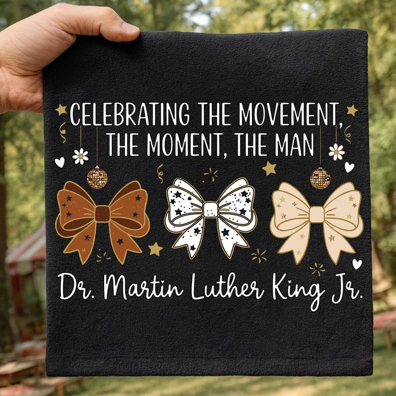 May include: Black towel with the text "CELEBRATING THE MOVEMENT, THE MOMENT, THE MAN" and "Dr. Martin Luther King Jr." in white. The design includes three bow ties in brown, white, and beige.