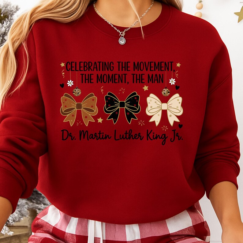 May include: A red sweatshirt with the text "CELEBRATING THE MOVEMENT, THE MOMENT, THE MAN" and "Dr. Martin Luther King Jr." printed on it. The design includes three decorative bows in gold, black, and white.