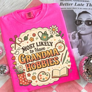 May include: A bright pink t-shirt with the phrase "MOST LIKELY to Have GRANDMA HOBBIES" in a retro design. The graphic includes illustrations of knitting needles, a watering can, flowers, and other craft-related items. The shirt is a Comfort Colors brand.