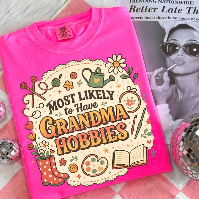 May include: A bright pink t-shirt with the phrase "MOST LIKELY to Have GRANDMA HOBBIES" in a retro design. The graphic includes illustrations of knitting needles, a watering can, flowers, and other craft-related items. The shirt is a Comfort Colors brand.