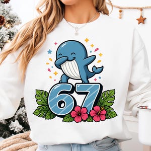 May include: White sweatshirt featuring a blue cartoon whale doing a dab pose. The number "67" is below the whale, decorated with tropical leaves and pink flowers. Colorful confetti and stars surround the whale.